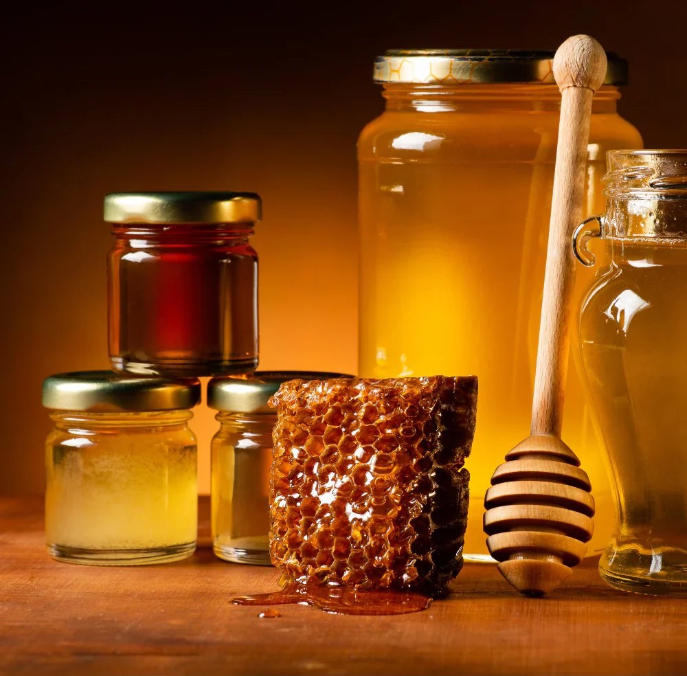 Jars of raw honey beside a dripping honeycomb and a wooden dipper